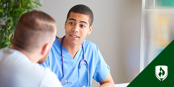 蓝刷男性医务工作者坐在办公桌前对模糊前景中的病人说话