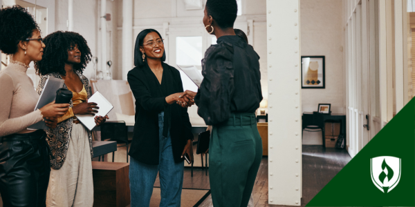 A female intern in a black blazer and turquoise pants shakes hands with a female employee in a group of people